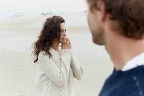 Woman with long brown hair with her hands to her face looking off into the distance, with a man in the distance looking at her