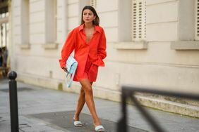 woman wearing an orange button-down shirt, orange shorts, and pastel blue mules